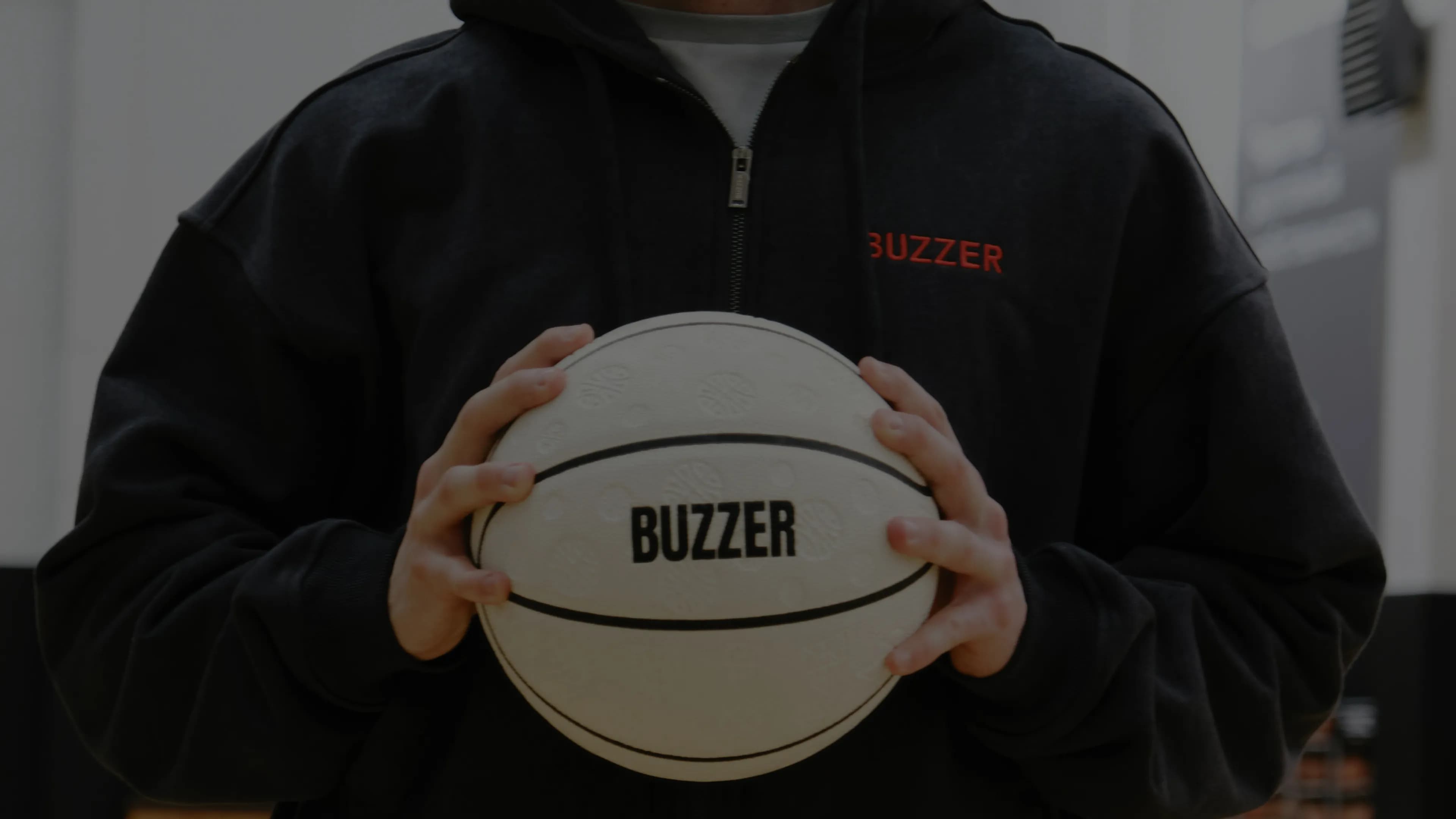Крупный план: в руках белый баскетбольный мяч BUZZER, на груди — вышивка на худи.